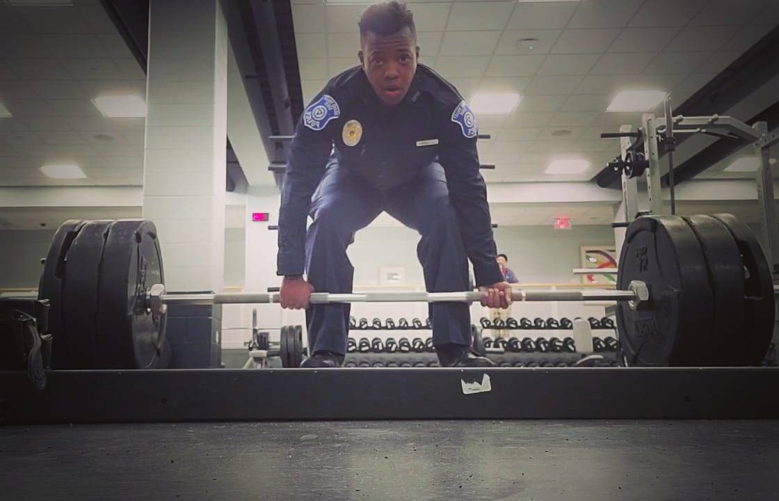 Julian Luebke dressed in his GVPD uniform, bending down to complete a deadlift with a straight bar and weights